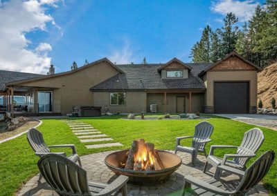 a wide shot of the Eaglecrest home's backyard, including a beautiful firepit and a brown, rustic angled home on a hill with pine forest mountains in the background