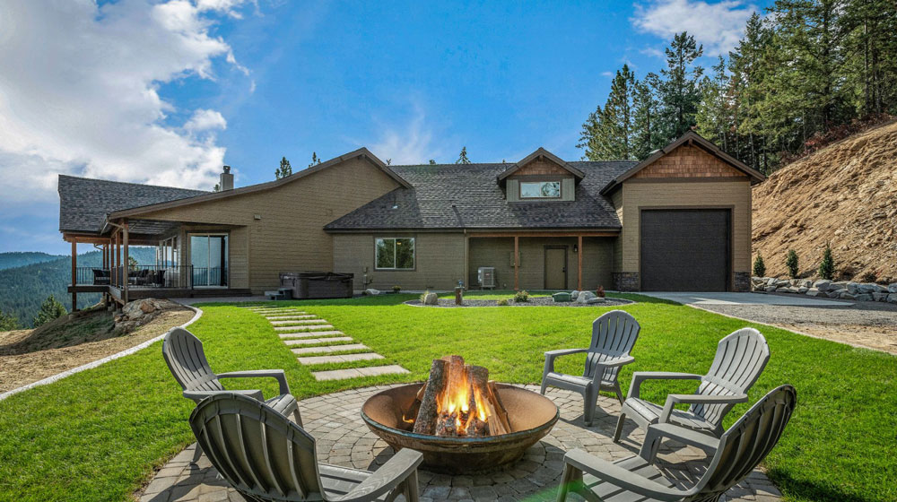 a wide shot of the Eaglecrest home's backyard, including a beautiful firepit and a brown, rustic angled home on a hill with pine forest mountains in the background
